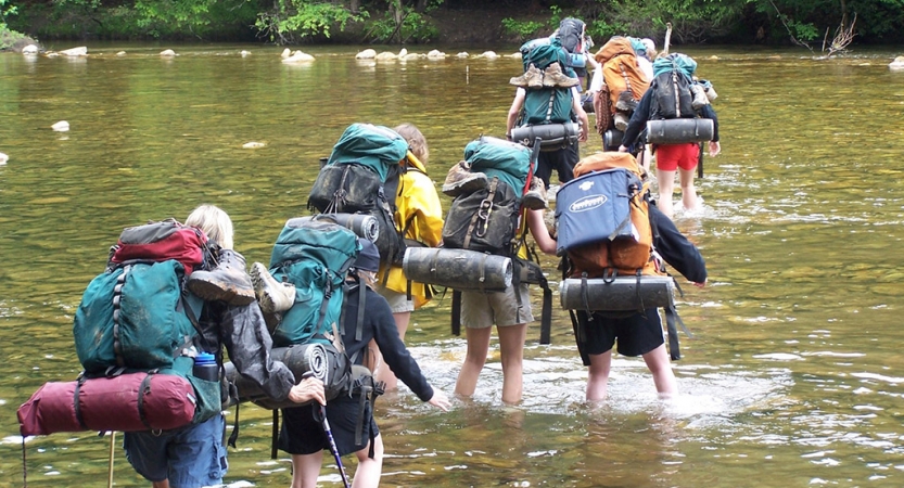 A group of people wearing backpacks cross a river in ankle-deep water. 